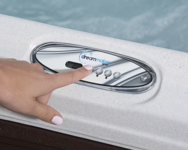 Close-up of a hand pressing a button on a DreamMaker spa control panel by a pool edge, showing a small digital display.