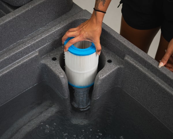 Person inserts a cylindrical pool/spa filter cartridge into a dark-gray filter housing, blue end cap visible, water nearby